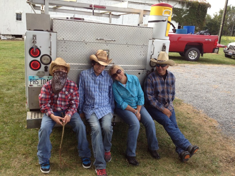 Three males with cowboy hats and one woman sit on the bumper of a fire truck